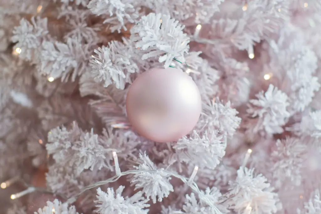 A matte finish light pink bauble hung on branches of a white artificial tree with white string lights