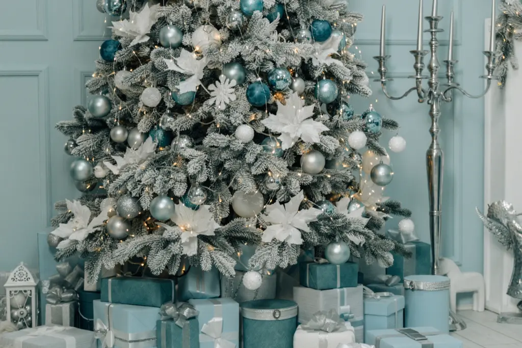 base of a blue and white decorated white artificial Christmas tree with wrapped blue presents around the base. Blue wall in background tall freestanding silver candle holder to the side