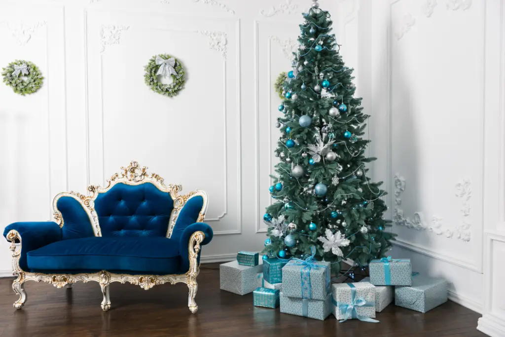 A white room with christmas tree in corner with blue decorations. Navy blue vintage style double arm chair next to it