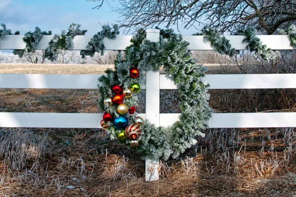 Hanging fir wreath with multi-colored baubles on left side on white fence, country backdrop